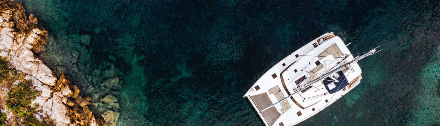 A drone view of a catamaran yacht anchored in a Mediterranean bay surrounded by dark emerald water