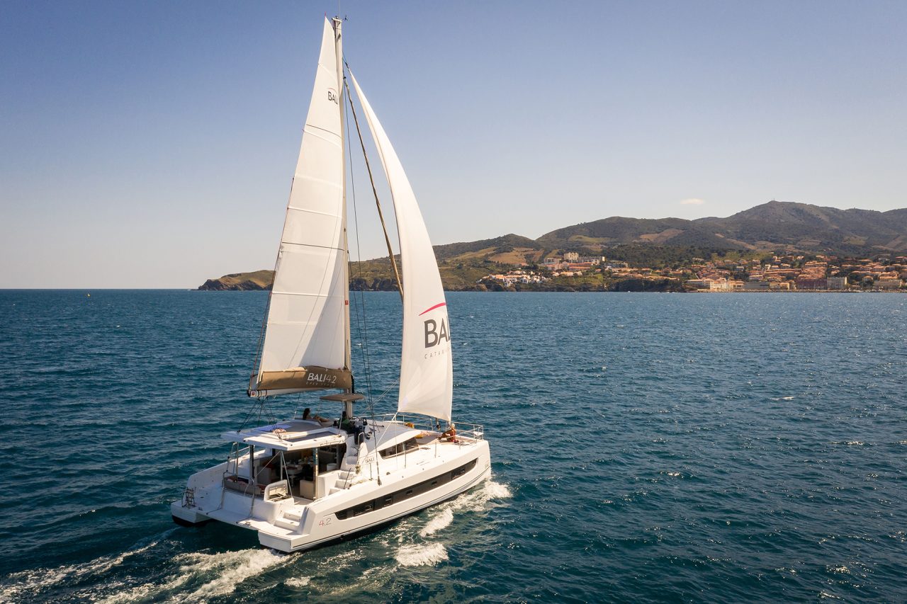 Bali 4.2 sailing near the coast with a man at the helm and women enjoying the foredeck and the flybridge