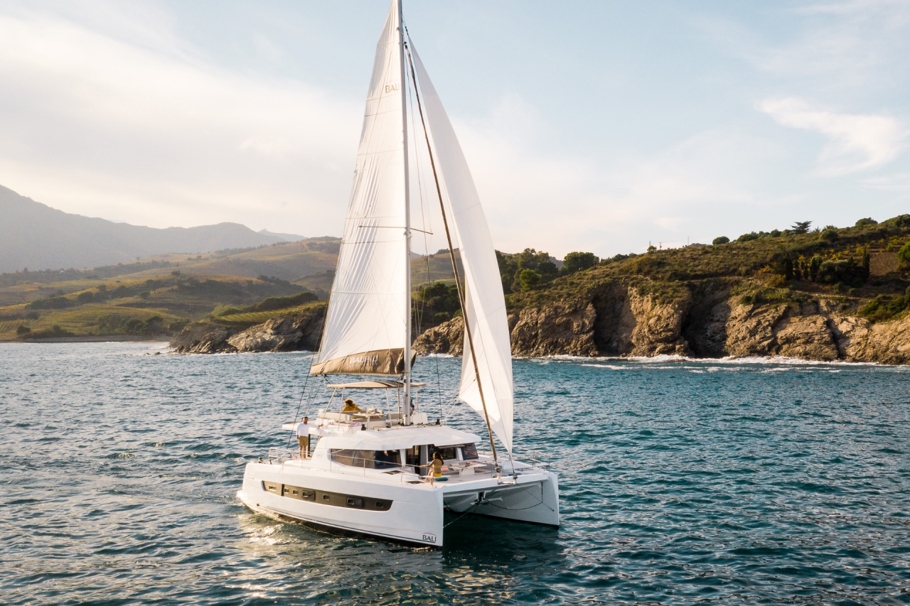 One of the popular Bali sailing catamarans sailing with a rugged coastal backdrop, with cliffs and blue water