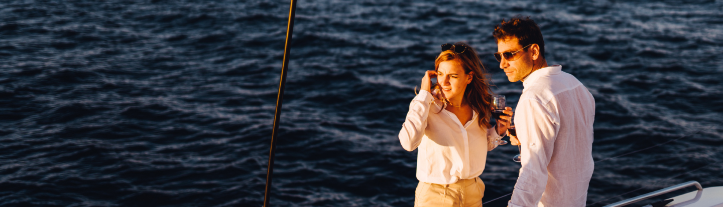 A couple on the side deck of a sailing catamaran at sunset, surrounded by deep blue water