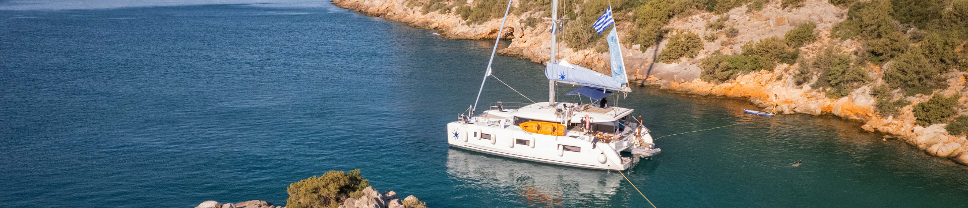 A sailing catamaran is anchored in a Mediterranean bay surrounded by rugged cliffs on both sides