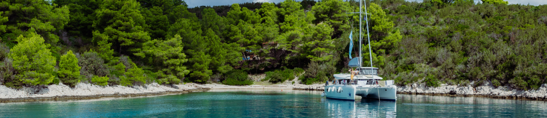 A sailing catamaran anchored in a Mediterranean bay in front of an island with lush greenery
