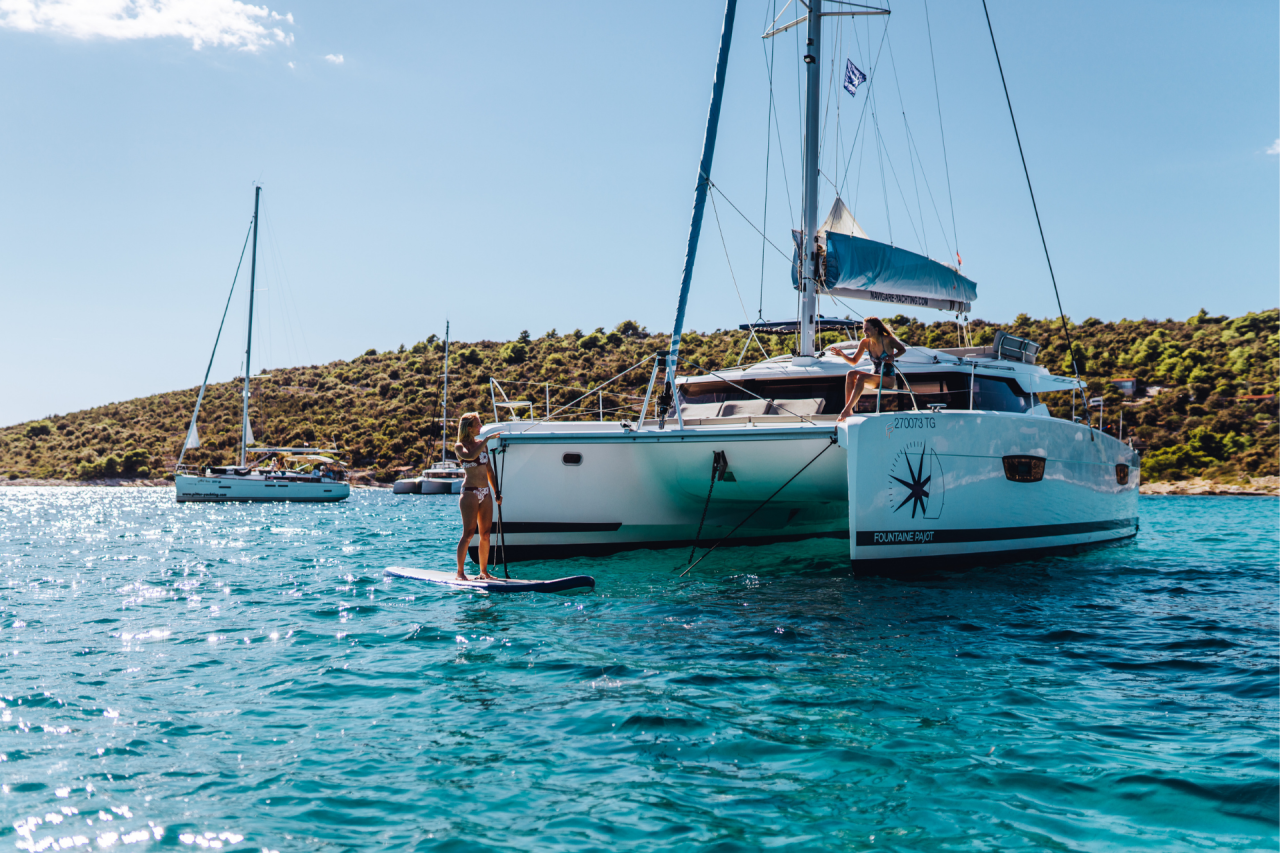 A sailing catamaran at anchor with a woman sitting on the foredeck and a woman on a paddleboard