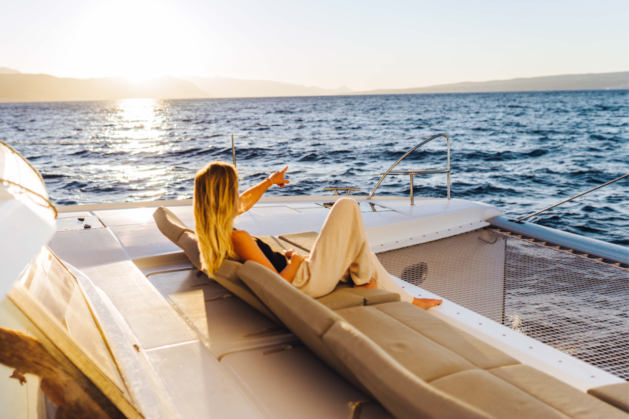 A woman on the forward sunbed of a sailing catamaran, pointing at the water, presumably at a dolphin
